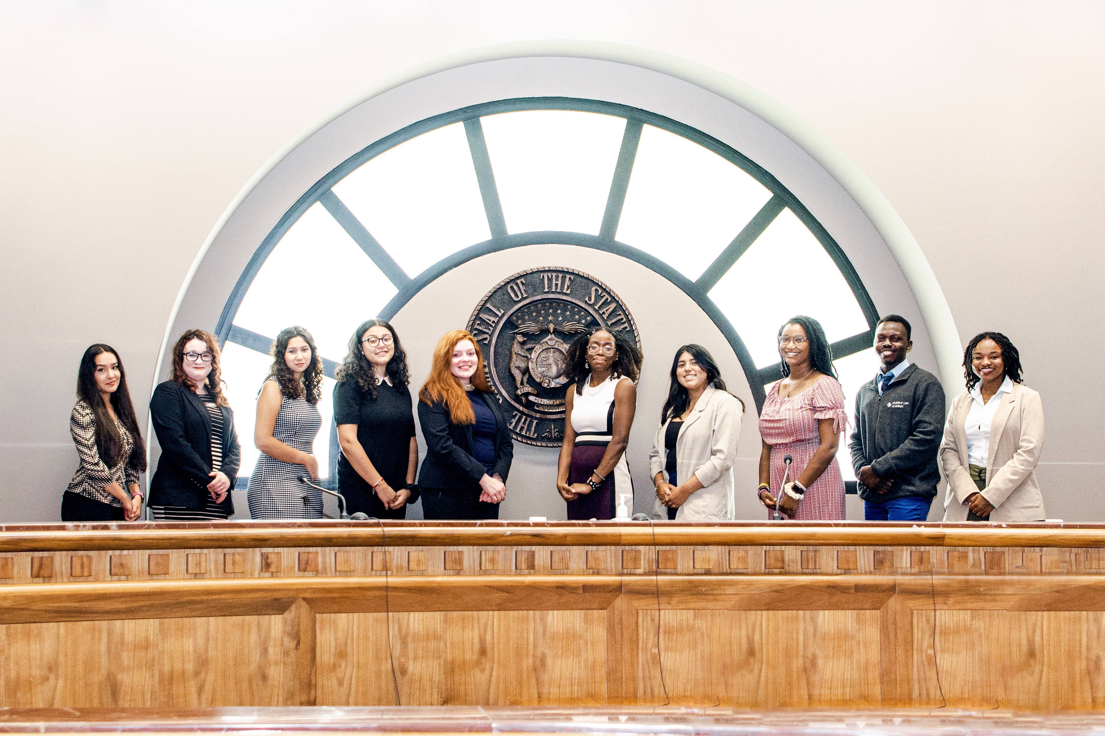 Image of 10 college students posing in front of a state seal and circular window.